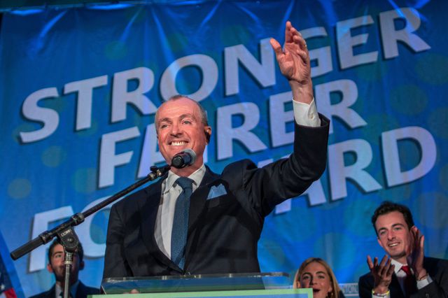 Phil Murphy raises his hand while on stage, in front of his "stronger fairer forward" slogan